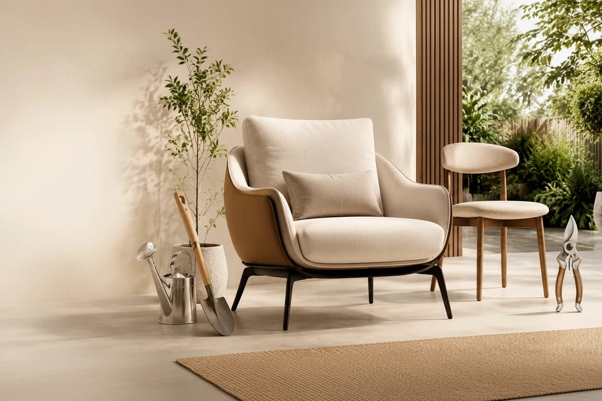 A styled interior scene showing a beige upholstered armchair with dark legs as the main product, alongside a matching side chair, garden tools, and outdoor greenery in the background. Large text on the left reads 'Create a Home You'll Love' with smaller descriptive copy and buttons labeled 'Shop Collection' and 'Explore Deals'.